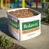 Rolawn play grade bark bulk bag sat on a concrete path with a colourful play area behind that has a play bark safety surface to protect children from falling